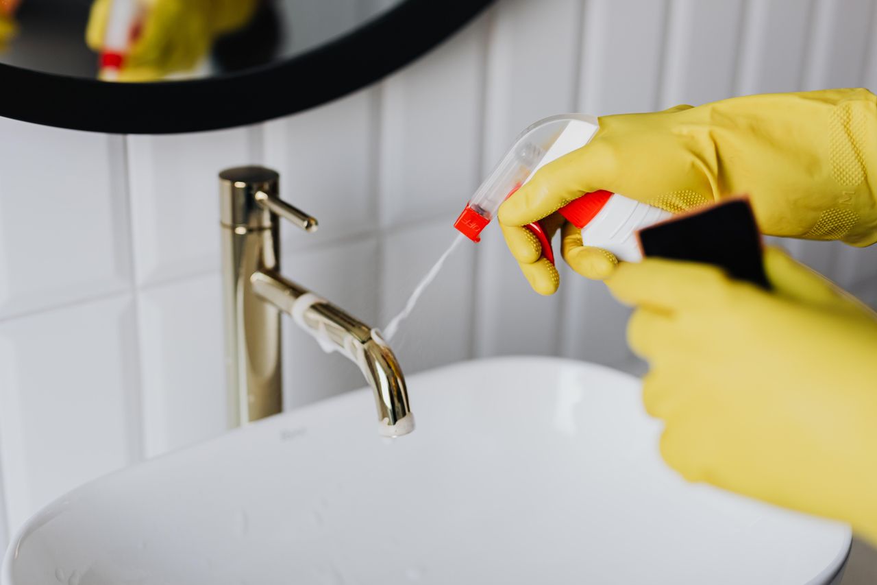 Vue depuis le dessus d’une femme de ménage anonyme portant des gants en caoutchouc jaunes et pulvérisant un produit nettoyant sur un robinet d’eau chromé dans une salle de bain